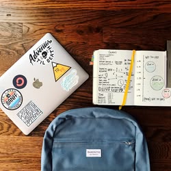 Ein Rucksack, ein MacBook mit Stickern und ein offenes Notizbuch liegen auf einem Tisch