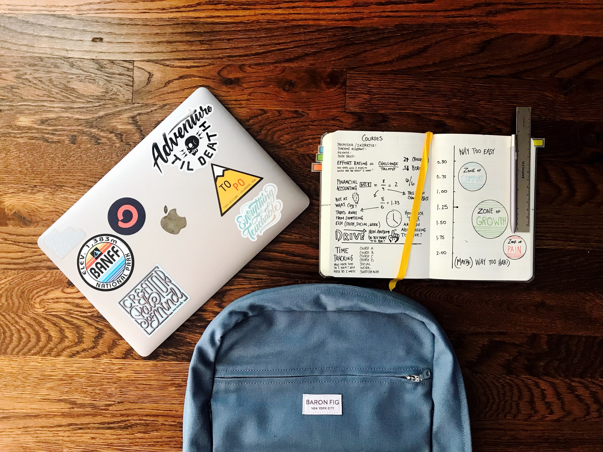 Ein Rucksack, ein MacBook mit Stickern und ein offenes Notizbuch liegen auf einem Tisch