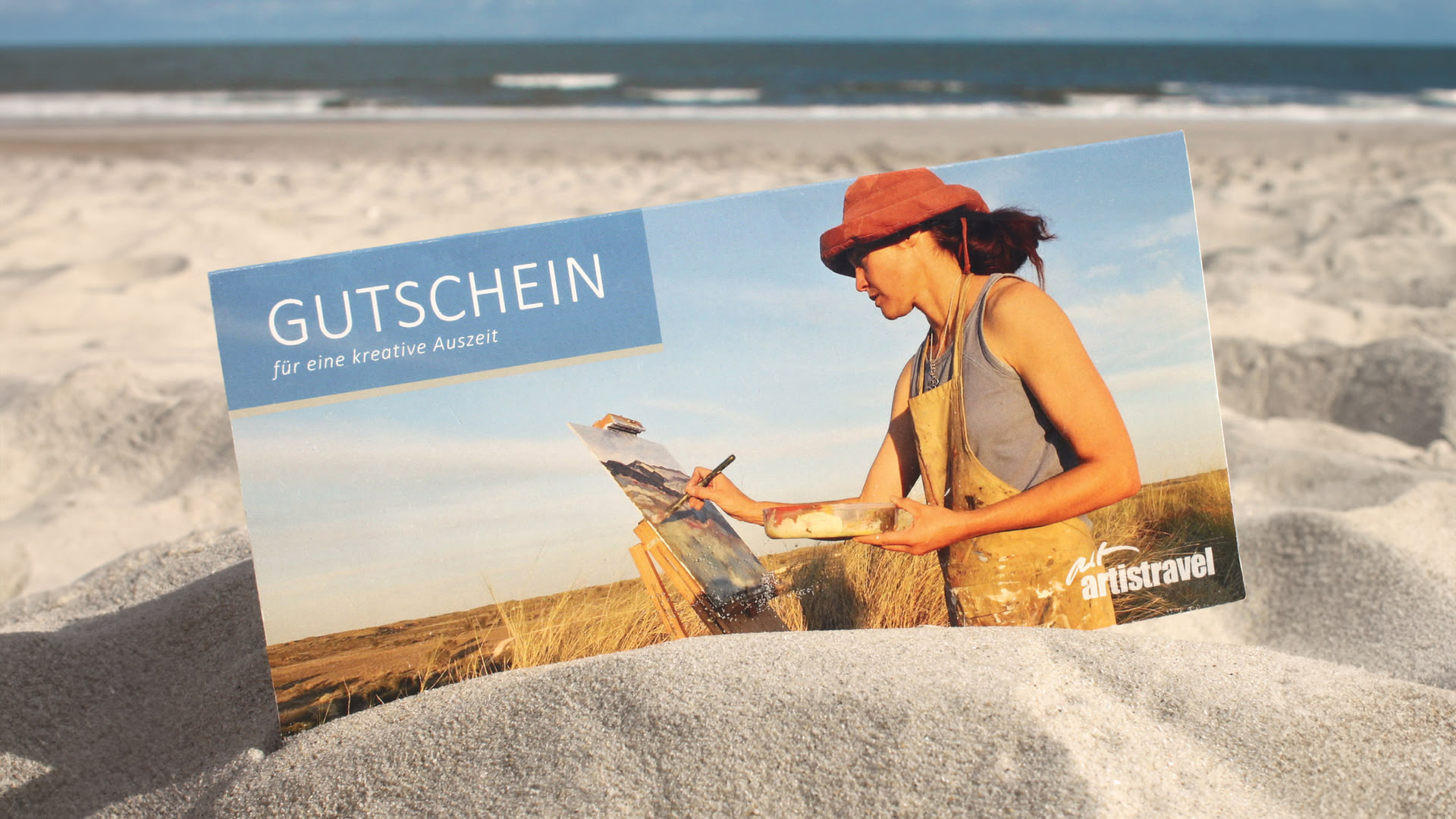 Ein artistravel-Gutschein steckt im Sand am Strand 