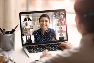 Blick über die Schulter einer Frau, auf ihrem Laptop ist eine Video-Konferenz geöffnet