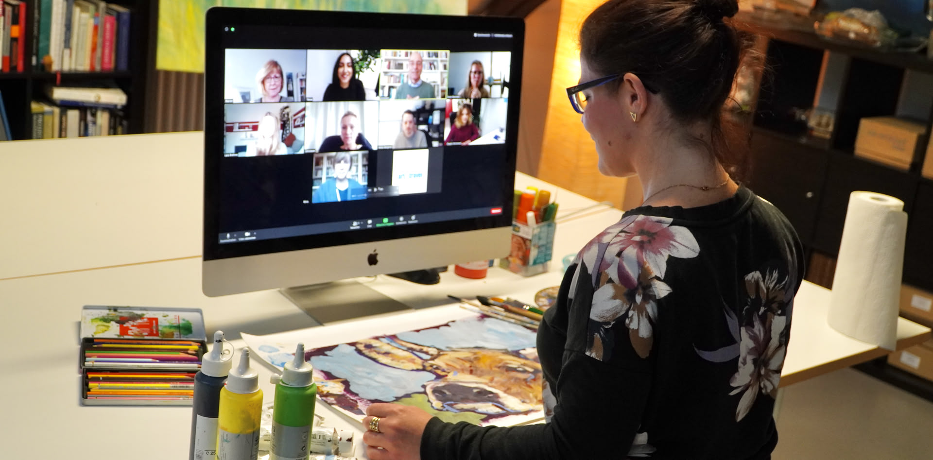 Eine Online-Kursteilnehmerin sitzt vor dem PC mit Zoom-Meeting, vor ihr liegt ihr gemaltes Bild