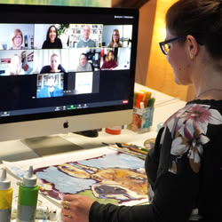 Eine Online-Kursteilnehmerin sitzt vor dem PC mit Zoom-Meeting, vor ihr liegt ihr gemaltes Bild