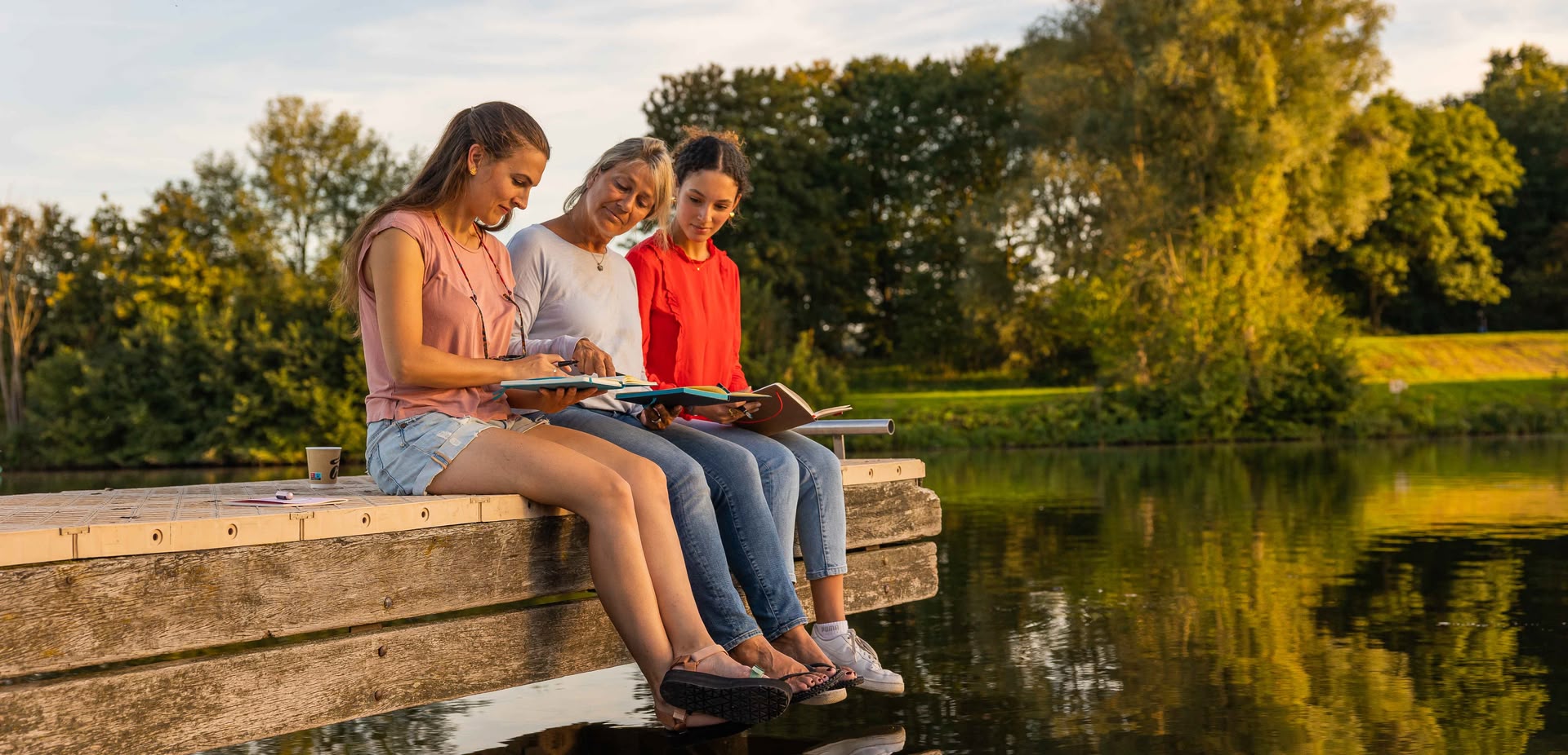 3 Frauen sitzen auf einem Steg am Wasser und schauen in ihre Notizbücher