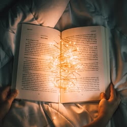Blick von oben auf ein aufgeschlagenes Buch mit einer Lichterkette darin, das von 2 Händen festgehalten wird und auf einem Bett liegt