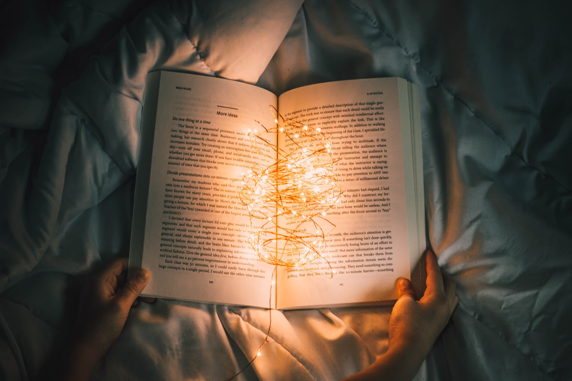 Blick von oben auf ein aufgeschlagenes Buch mit einer Lichterkette darin, das von 2 Händen festgehalten wird und auf einem Bett liegt
